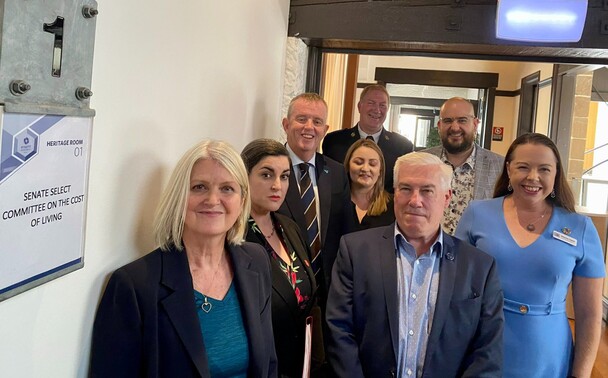 A group of adults including four women and four men, all from Uniting Vic.Tas and Wesley Mission, wait in a corridor to give evidence at the Senate Cost of Living Inquiry. A sign on the wall on the left reads 'Senate Select Committee on the Cost of Living'. 
