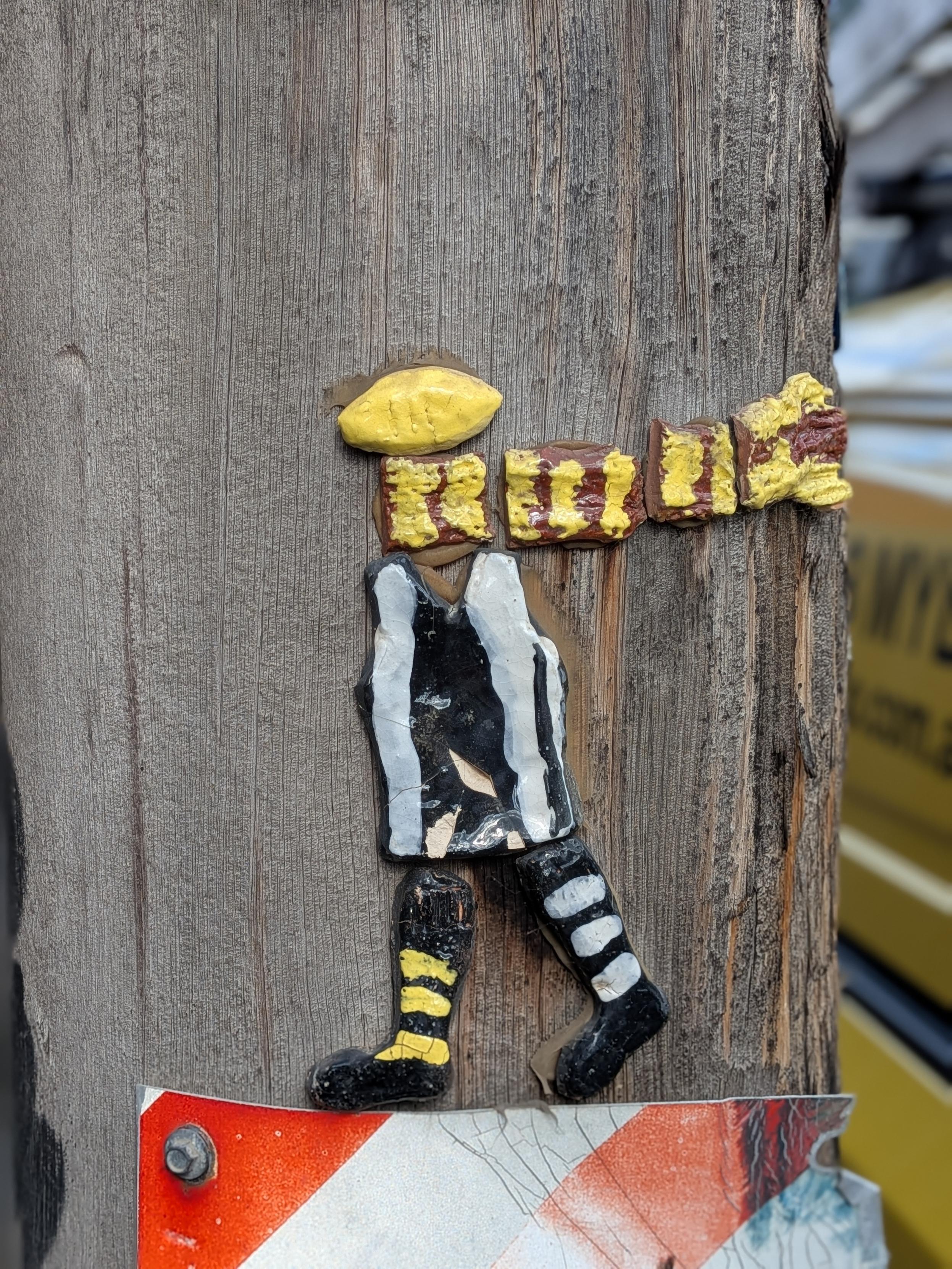 A clay footy player glued to a wooden electricity pole. Its pretty cute, has an Aussie footy for s head, but, it can't decide who to support, wearing a black and white jumper with a brown and yellow scarf.