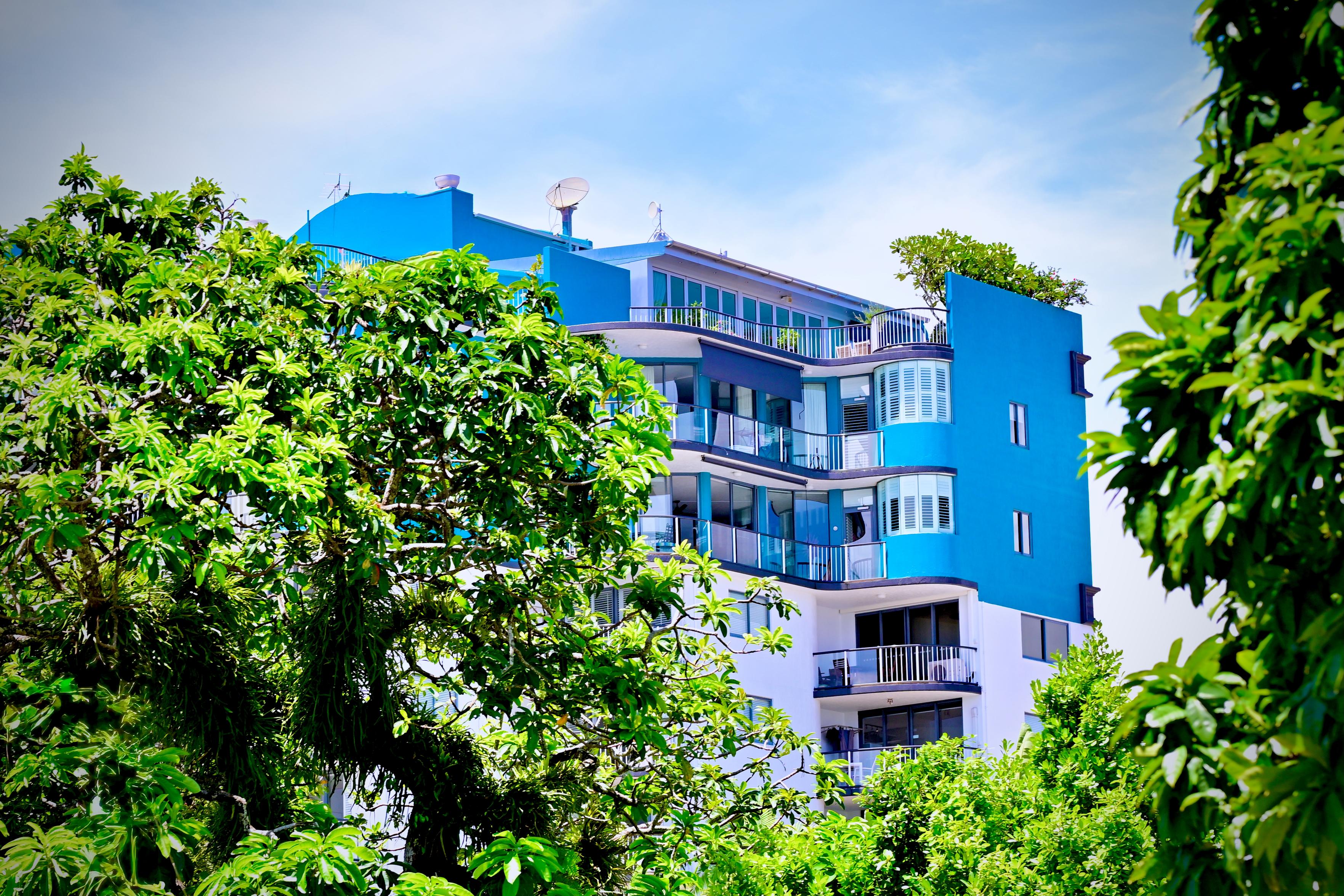 A multi-storey residential tower in blue and white, half-swallowed by the canopy in front of it. Curved balconies and louvred shutters run up the facade, with rooftop greenery spilling over the upper levels. The trees push in hard from both sides, making the building feel less like a city block and more like something the tropics are quietly reclaiming.