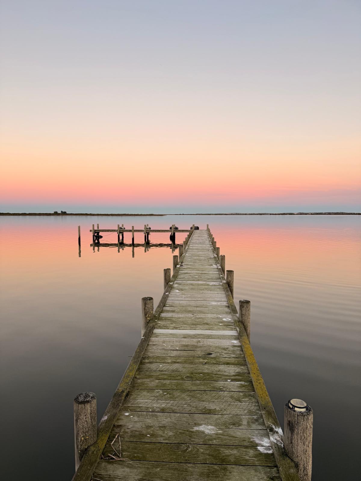 Wooden dock over lake. There’s a vibrant pink sunset and is reflected back on the lake.
